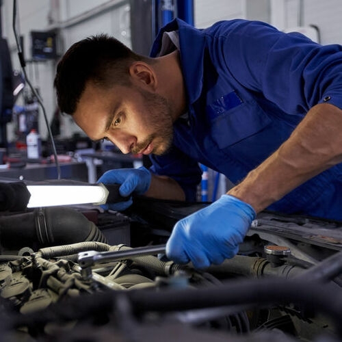 car service, repair, maintenance and people concept - auto mechanic man with lamp working at workshop